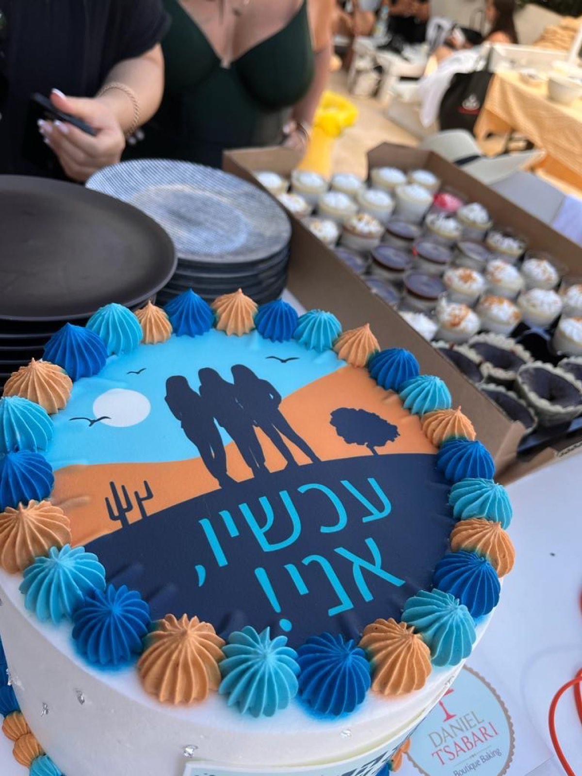 עוגה מעוצבת עם איור הלוגו של עכשיו, אני! מוקפת קישוטי קרם כחולים וכתומים